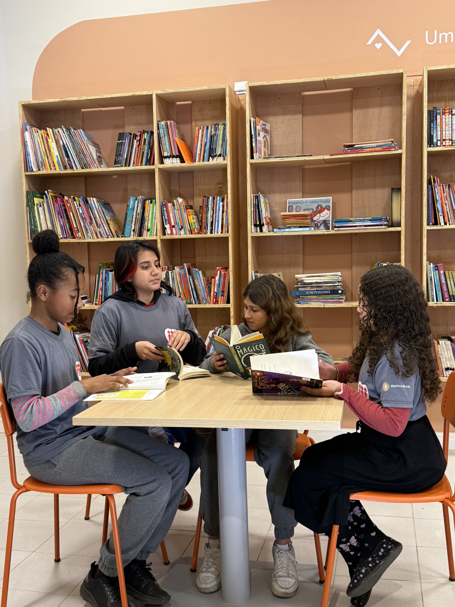 Quatro jovens estudantes sentadas ao redor de uma mesa em uma biblioteca, engajadas na leitura e na troca de ideias. A cena, com estantes repletas de livros ao fundo, transmite um ambiente de concentração, colaboração e paixão pelo aprendizado.